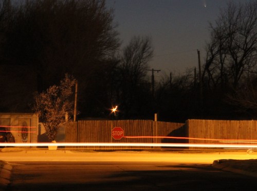 A nighttime scene featuring a stop sign, a wooden fence, and light trails from passing vehicles.