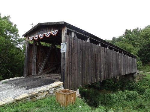 A wooden covered bridge with a stone foundation, decorated with red, white, and blue bunting, surrounded by greenery.