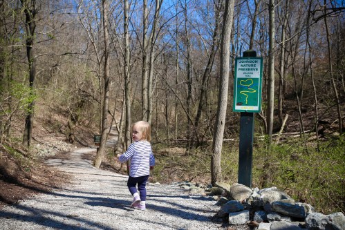 A toddler walks along a gravel path in a wooded area, with a sign for Johnson Nature Preserve in the background.