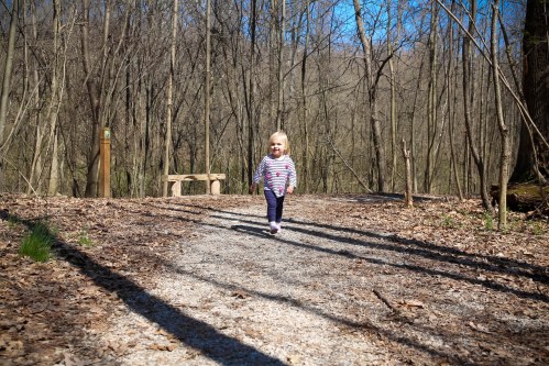 A young child with blonde hair walks happily along a gravel path in a wooded area on a sunny day.