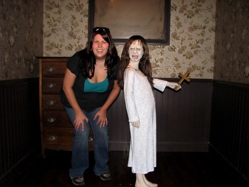 A woman smiles next to a creepy doll in a vintage room, holding a cross and wearing a nightgown.