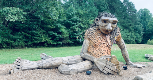 A large wooden sculpture of a seated figure with a natural setting of trees in the background.