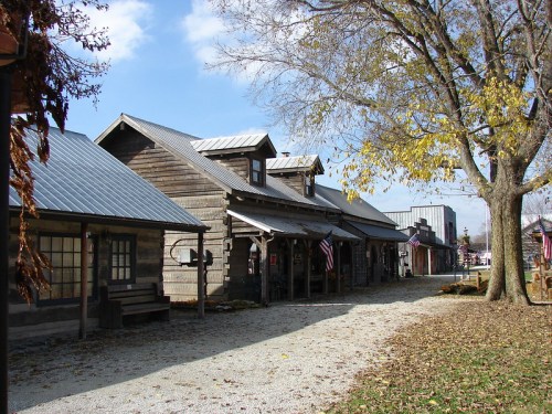 A rustic street scene featuring wooden buildings, a gravel path, and trees with autumn leaves under a blue sky.