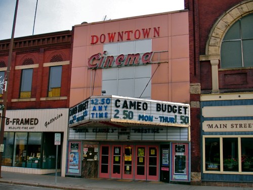 Historic cinema building with a marquee displaying movie titles and prices, surrounded by brick storefronts.