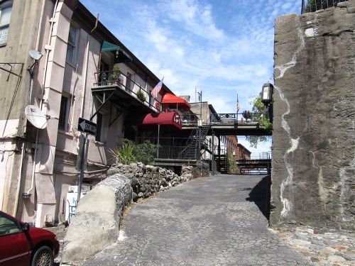 Narrow street with buildings, balconies, and a red awning, featuring a stone wall and a clear blue sky.