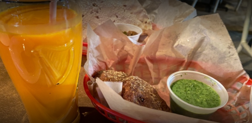 A glass of orange drink beside a basket with two rolls and a cup of green sauce on a table.