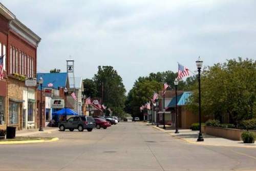 A quiet downtown street lined with American flags, featuring shops and trees under a cloudy sky.
