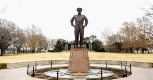 A tall bronze statue of a man stands on a pedestal in a park, surrounded by trees and a grassy area.