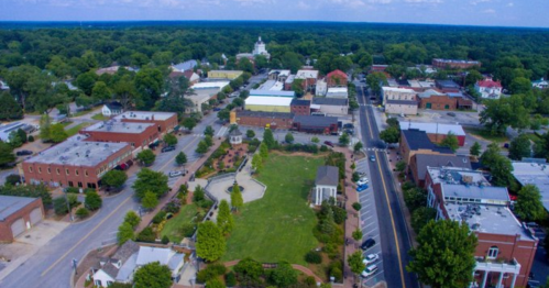 Aerial view of a small town with green spaces, buildings, and a central park area surrounded by trees.