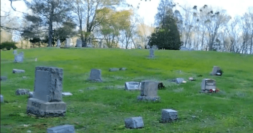 A peaceful cemetery with scattered gravestones on a grassy hillside, surrounded by trees and a clear sky.