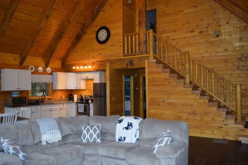 Cozy wooden cabin interior featuring a living area, kitchen, and staircase, with a large sectional sofa and rustic decor.