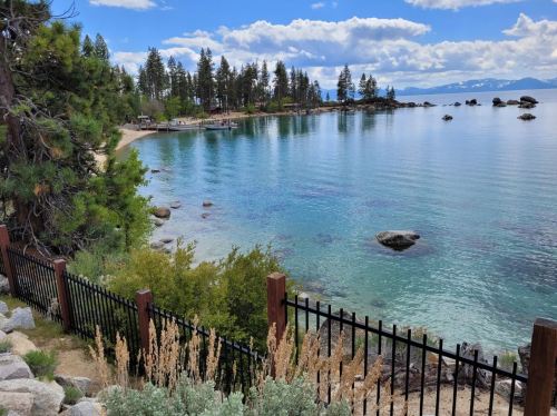 A serene lakeside view with clear water, rocky shores, and trees under a blue sky with fluffy clouds.