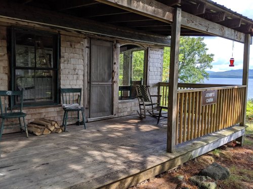 A rustic cabin porch with wooden chairs, a sign reading "Jenny's Cabin," and a view of a lake and trees.