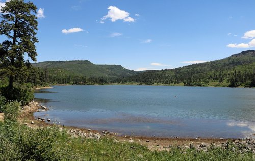 A serene lake surrounded by green hills and trees under a clear blue sky with a few clouds.