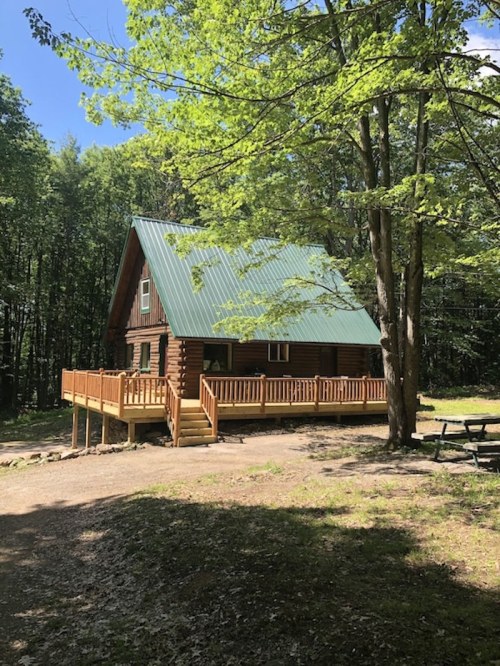 A rustic log cabin with a green metal roof, surrounded by trees and a gravel path, featuring a wooden deck.