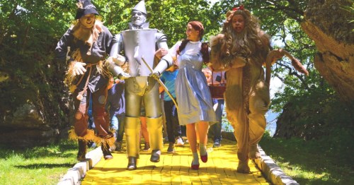 A group dressed as characters from "The Wizard of Oz" walks along a yellow brick road in a lush, green setting.