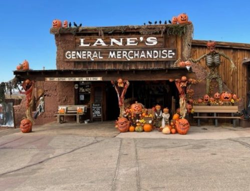 A rustic general store decorated with pumpkins, skeletons, and Halloween decor, set against a clear blue sky.