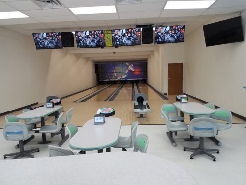 A bowling alley interior featuring empty lanes, screens, and tables with chairs. Bright and spacious atmosphere.