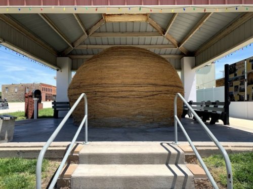 A large, round straw sculpture sits under a pavilion, with steps and benches visible in the foreground.