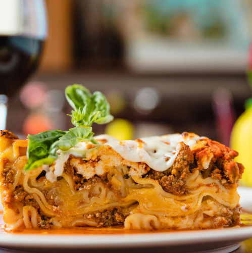 A slice of lasagna with layers of meat, cheese, and sauce, garnished with a basil leaf, next to a glass of red wine.