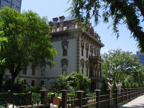 A grand, historic mansion surrounded by lush greenery and a decorative fence on a sunny day.