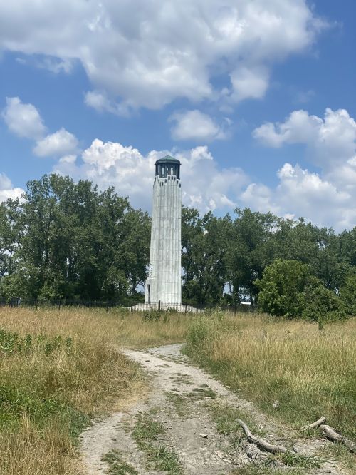 A tall white lighthouse stands amidst green trees under a blue sky with fluffy clouds, surrounded by tall grass.