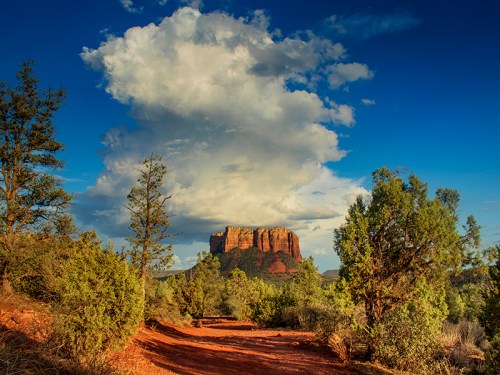 A scenic view of a red rock formation under a blue sky with fluffy clouds, surrounded by green shrubs and trees.