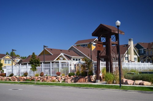 A residential community entrance with a wooden archway, white fence, and colorful houses under a clear blue sky.