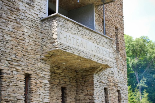 Close-up of a stone building with a sign reading "Château La Roche" on a balcony. Lush greenery in the background.