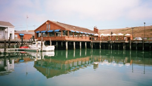 A wooden restaurant on a pier, reflecting in calm water, with boats and umbrellas in the background.