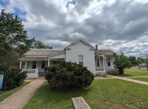 A white, single-story house with a porch, surrounded by greenery and under a cloudy sky.