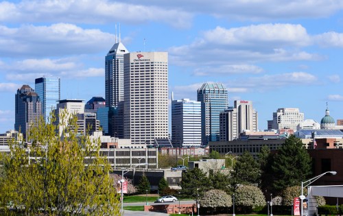 A city skyline featuring tall buildings and a blue sky with clouds, surrounded by greenery in the foreground.