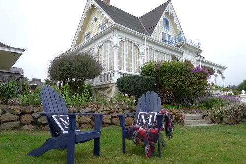 Two blue Adirondack chairs with blankets sit on a lawn in front of a large, elegant house surrounded by greenery.