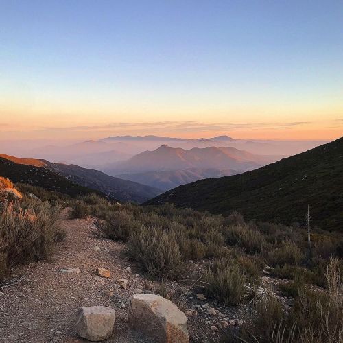 A scenic mountain landscape at sunset, with rolling hills and a path lined with shrubs in the foreground.