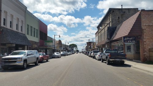 A view of a small town street lined with shops and parked cars under a partly cloudy sky.