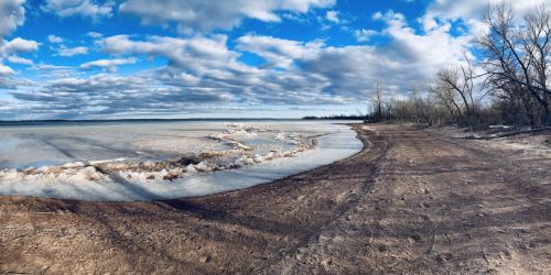 A serene lakeshore with ice patches, sandy beach, and cloudy blue sky, surrounded by bare trees.