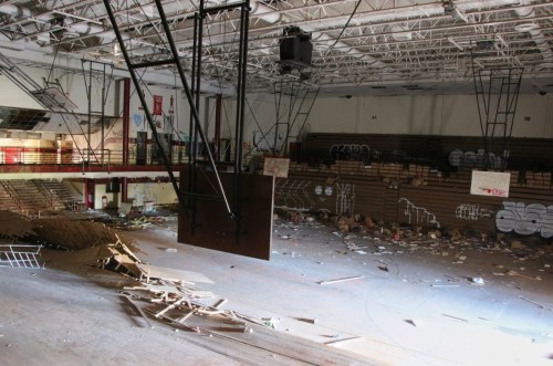 Abandoned gymnasium with debris, graffiti, and empty bleachers under a high ceiling.