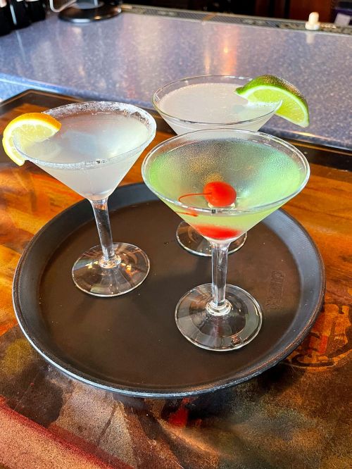 A tray with four colorful cocktails, including a lime garnish and cherries, on a blue countertop.