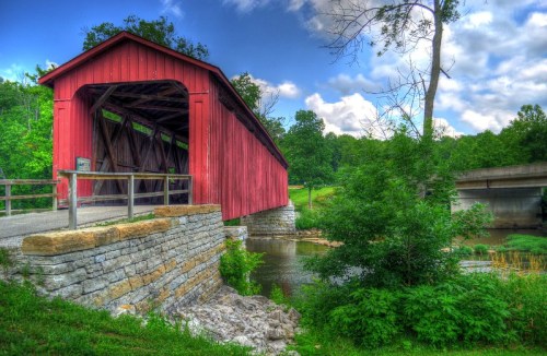 A red covered bridge spans a river, surrounded by lush greenery and a partly cloudy sky.