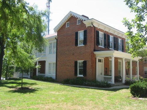 Two-story brick house with white trim, black shutters, and a porch, surrounded by green grass and trees.