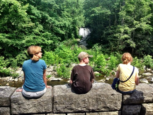 Three people sit on a stone wall, overlooking a lush green landscape and a waterfall in the distance.