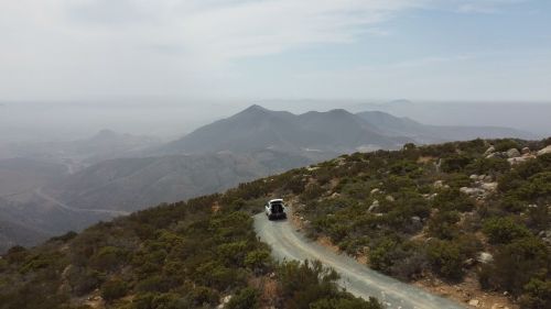 Aerial view of a winding dirt road through hilly terrain, with a vehicle driving towards distant mountains under a cloudy sky.