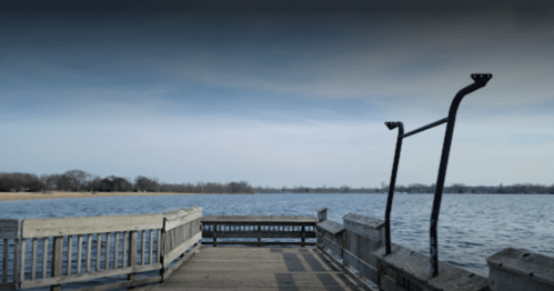 A wooden pier extends over a calm lake under a cloudy sky, with trees visible in the background.