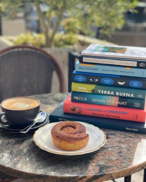 A coffee cup and a cinnamon roll on a table, with a stack of colorful books in the background.