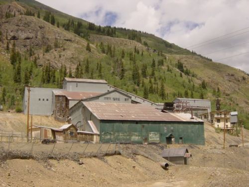 Abandoned industrial buildings nestled in a mountainous landscape, surrounded by trees and rocky terrain.