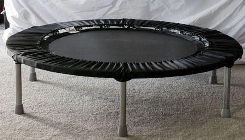 A round black trampoline with a padded edge, resting on four metal legs on a carpeted surface.