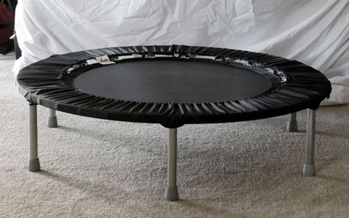 A round black trampoline with a padded edge, resting on metal legs, set against a light-colored background.