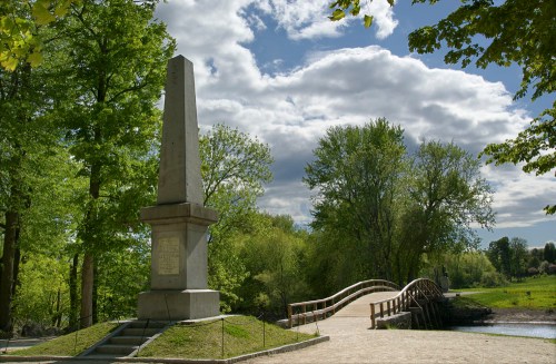 A stone obelisk stands near a wooden bridge, surrounded by lush green trees and a cloudy sky.