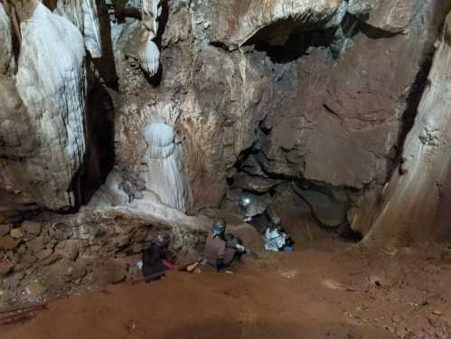 Two people sit on the ground inside a cave, surrounded by rock formations and dim lighting.