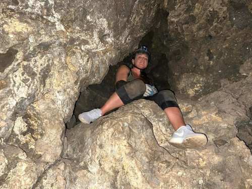A person sitting in a rocky cave, wearing a headlamp and climbing gear, with legs bent and hands resting on their knees.